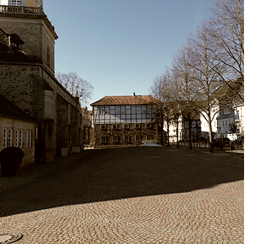 Zwischenstopp auf dem Platz neben der Kirche in Rinteln bei strahlendem Sonnenschein. Links vom Platz steht die Kirche, im Hintergrund ein Fachwerkhaus und rechts eine Reige kahler Bäume.